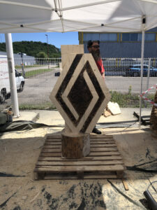 Totem publicitaire en bois sculpté pour Renault, avec le nom de la marque gravé verticalement et une petite voiture en bois au sommet.