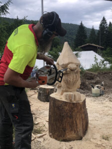 Vue de profil du sculpteur Sébastien Steffen, vêtu d'un t-shirt jaune et rouge, utilisant une tronçonneuse pour sculpter les traits d'un gnome de jardin assis sur une souche. Copeaux de bois volants.