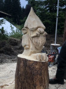 Sculpture sur bois d'un nain de jardin détaillée, avec une longue barbe et un bonnet pointu, par Sébastien Steffen. Vue en extérieur.