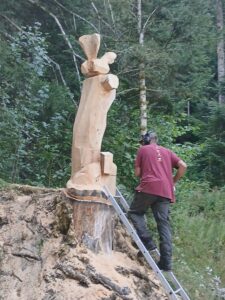 Début de la sculpture monumentale sur un tronc d'arbre perché sur un talus, avec l'artiste sur une échelle.