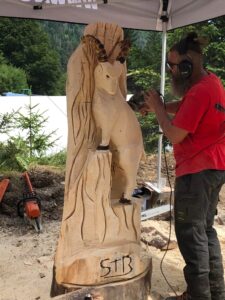 Sculpture sur bois d'un personnage assis, avec de longs cheveux et une barbe, œuvre de Sébastien Steffen. Assis sur un rondin de bois.