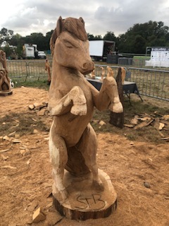 Une sculpture en bois grandeur nature d'un cheval cabré, vue de face, exposée sur un terrain jonché de copeaux de bois. L'arrière-plan montre un festival en plein air avec des tentes, des véhicules et des spectateurs. Sculpté par Sebastien Steffen
